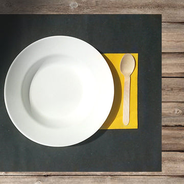 Tovaglietta carta paglia nera con tovagliolo di carta giallo, cucchiaio di legno e piatto bianco